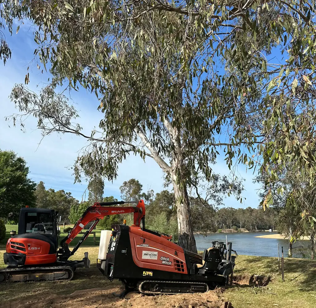 An orange excavator and drilling machine work on a grassy area beneath tall eucalyptus trees near a peaceful river, with blue sky and green foliage in the background.