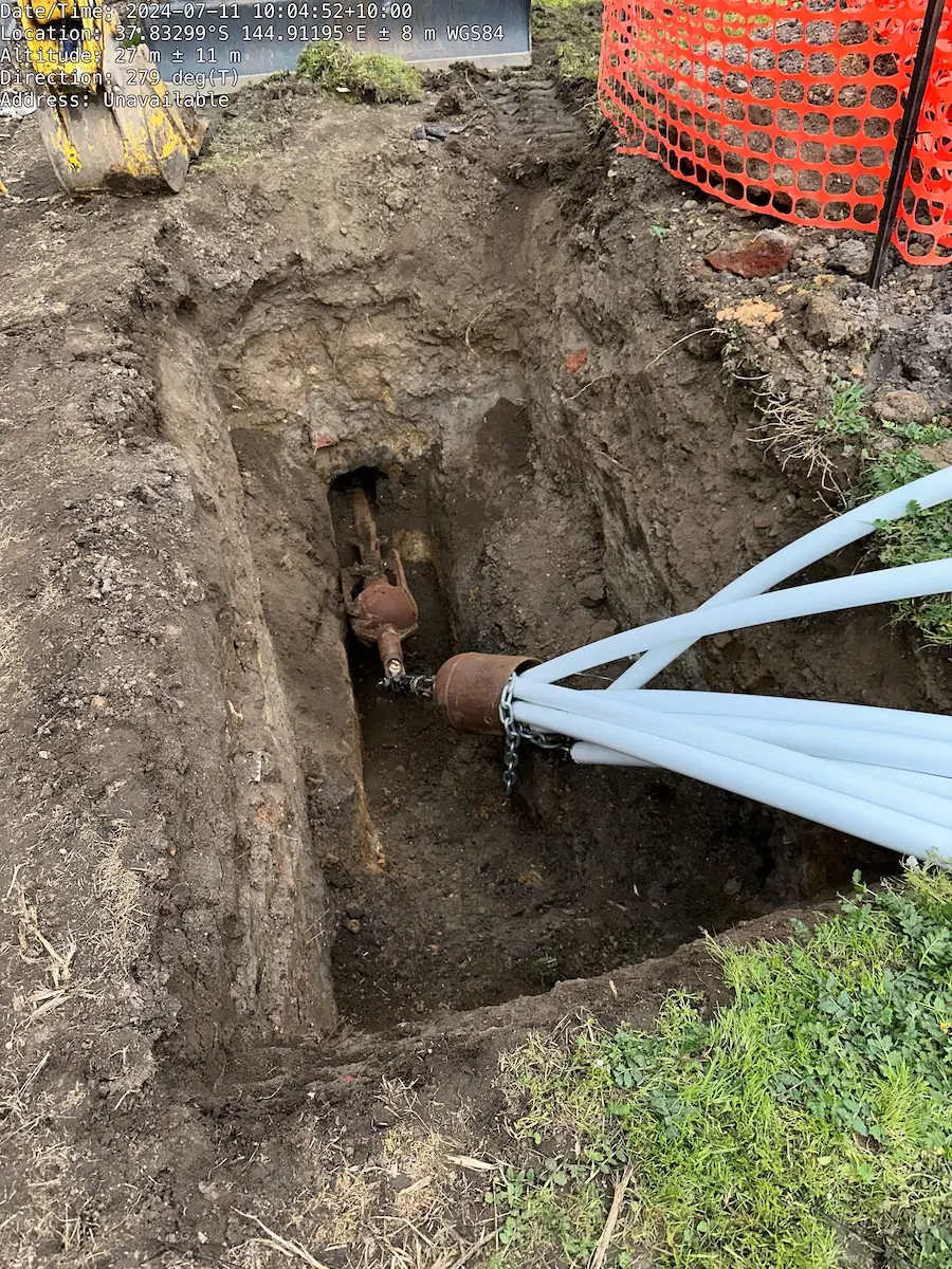 A deep trench in the ground exposes a buried valve connected to several white cables or pipes, with orange safety fencing and construction equipment visible at the edge of the site.
