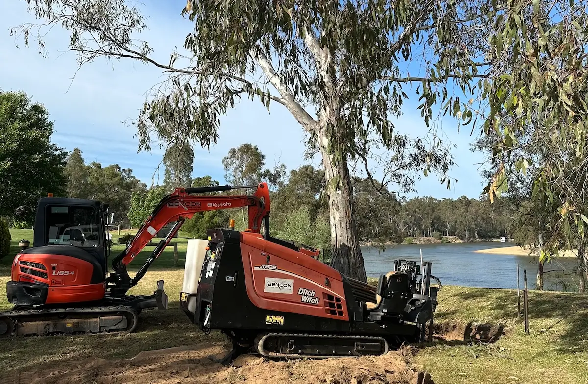 An orange drilling machine and a small excavator are working on grassy ground near a river, surrounded by trees under a clear blue sky.