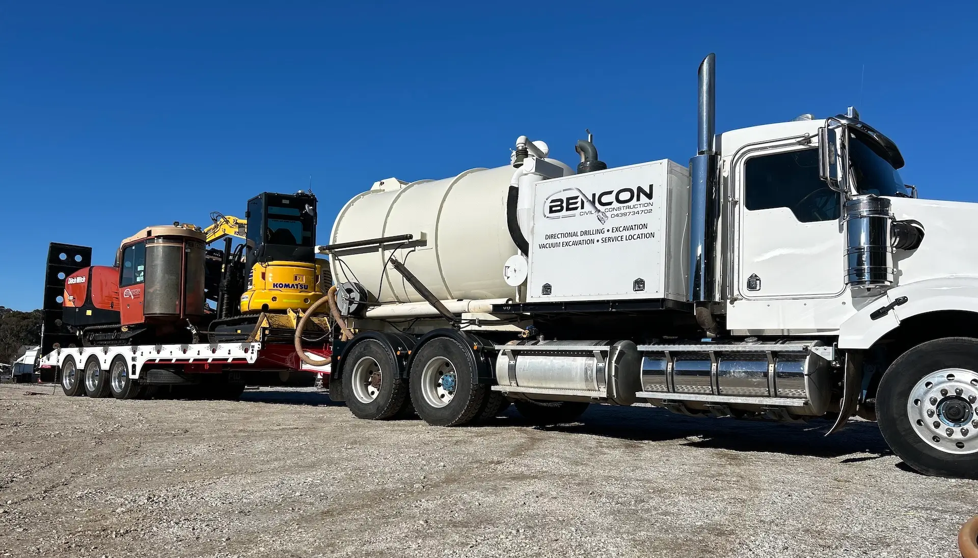 A white truck with a large tank on its back, labeled "BENCON," is parked on gravel, towing a flatbed trailer carrying construction equipment under a clear blue sky.