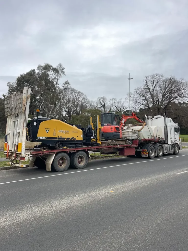 A flatbed truck parked on the roadside carries construction equipment, including a yellow Vermeer machine, a red mini excavator, and other machinery, with trees and a cloudy sky in the background.