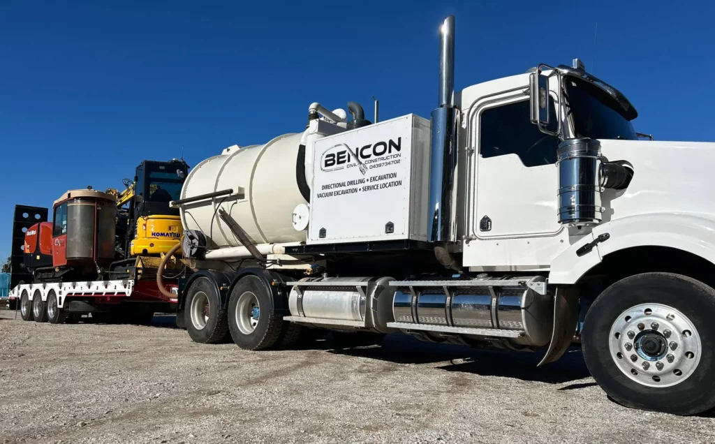 A white Bencon industrial vacuum truck, part of civil construction services, is parked on gravel, towing a flatbed trailer loaded with heavy construction equipment under a clear blue sky.