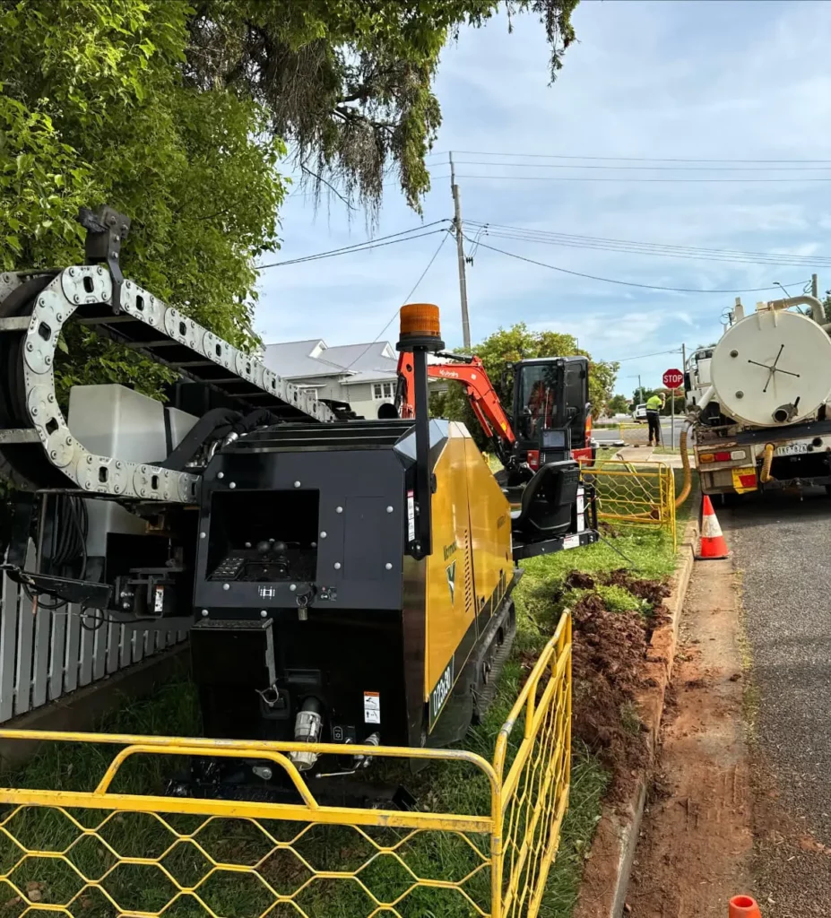A yellow horizontal drilling machine, used for civil construction services, is set up on a grassy roadside next to a fenced area, with construction cones and a truck nearby. Houses, utility poles, and trees stand in the background under a cloudy sky.