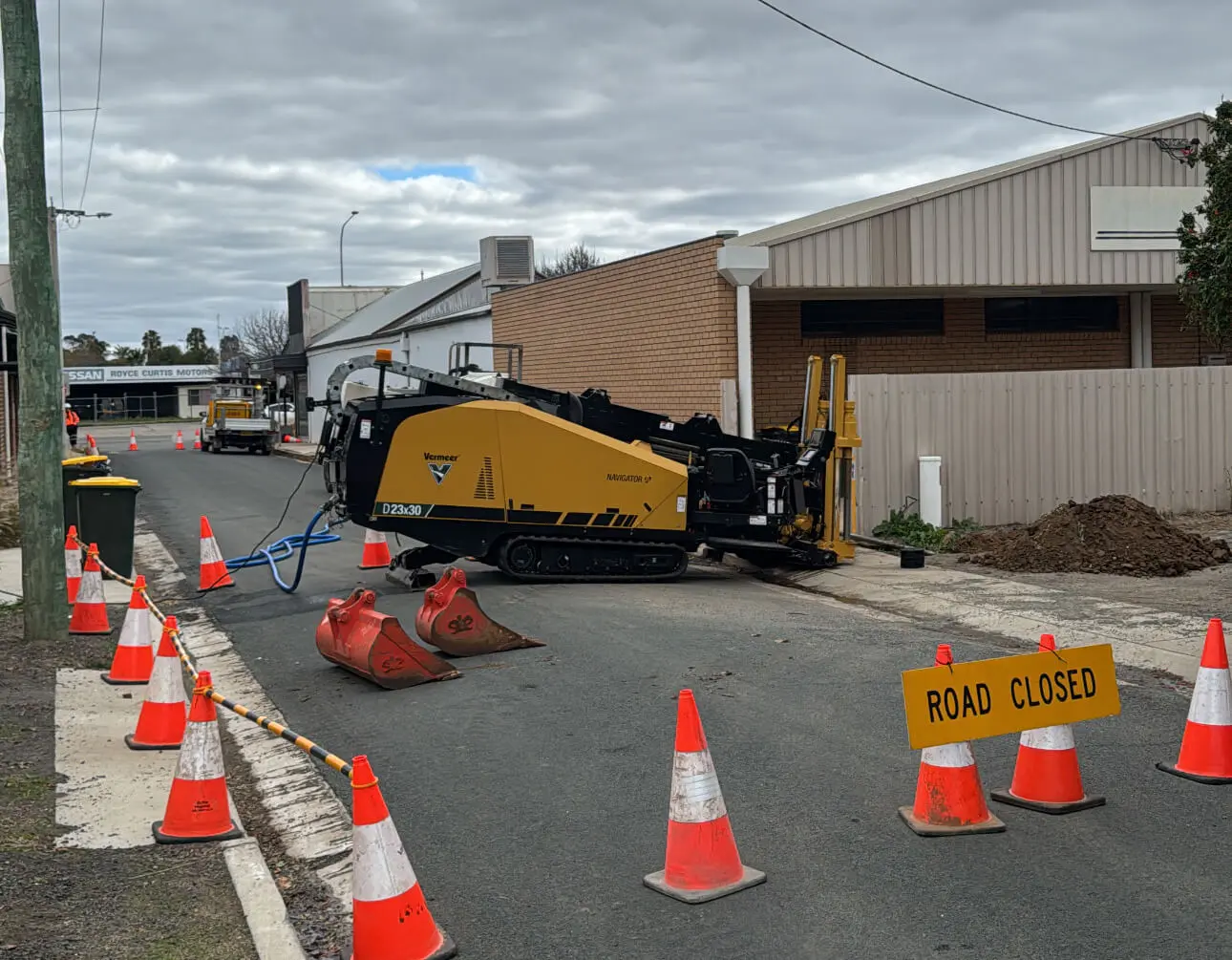 A civil construction company’s machine and equipment are positioned on a blocked road, surrounded by orange traffic cones. A "Road Closed" sign is visible, and the ground is dug up near a building under a cloudy sky.