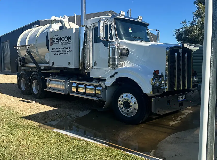 A large white vacuum truck with "BENCON" branding, part of a directional drilling company, is parked on a driveway near a building under a clear blue sky. The truck features shiny chrome accents and a cylindrical tank on its back.