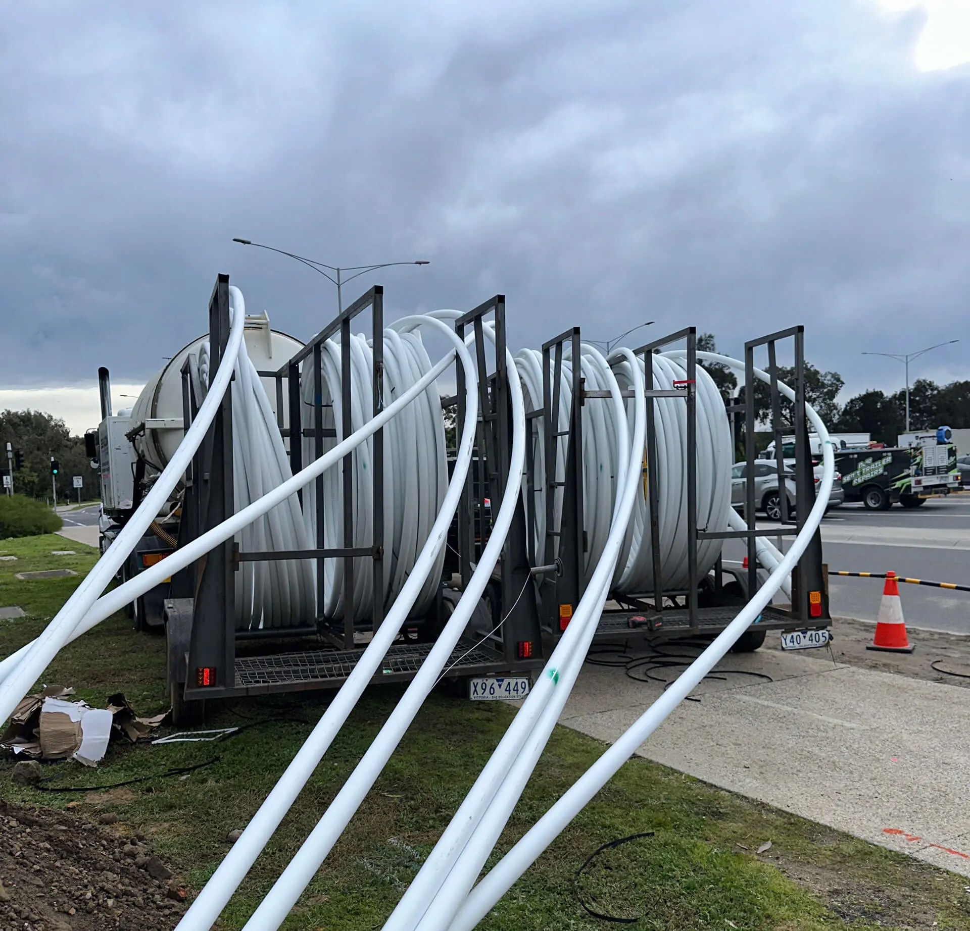 Large coils of white plastic piping, used by a directional drilling company, are mounted on a trailer next to a road, with several pipes extended outward. Traffic cones and vehicles are visible in the background under a cloudy sky.