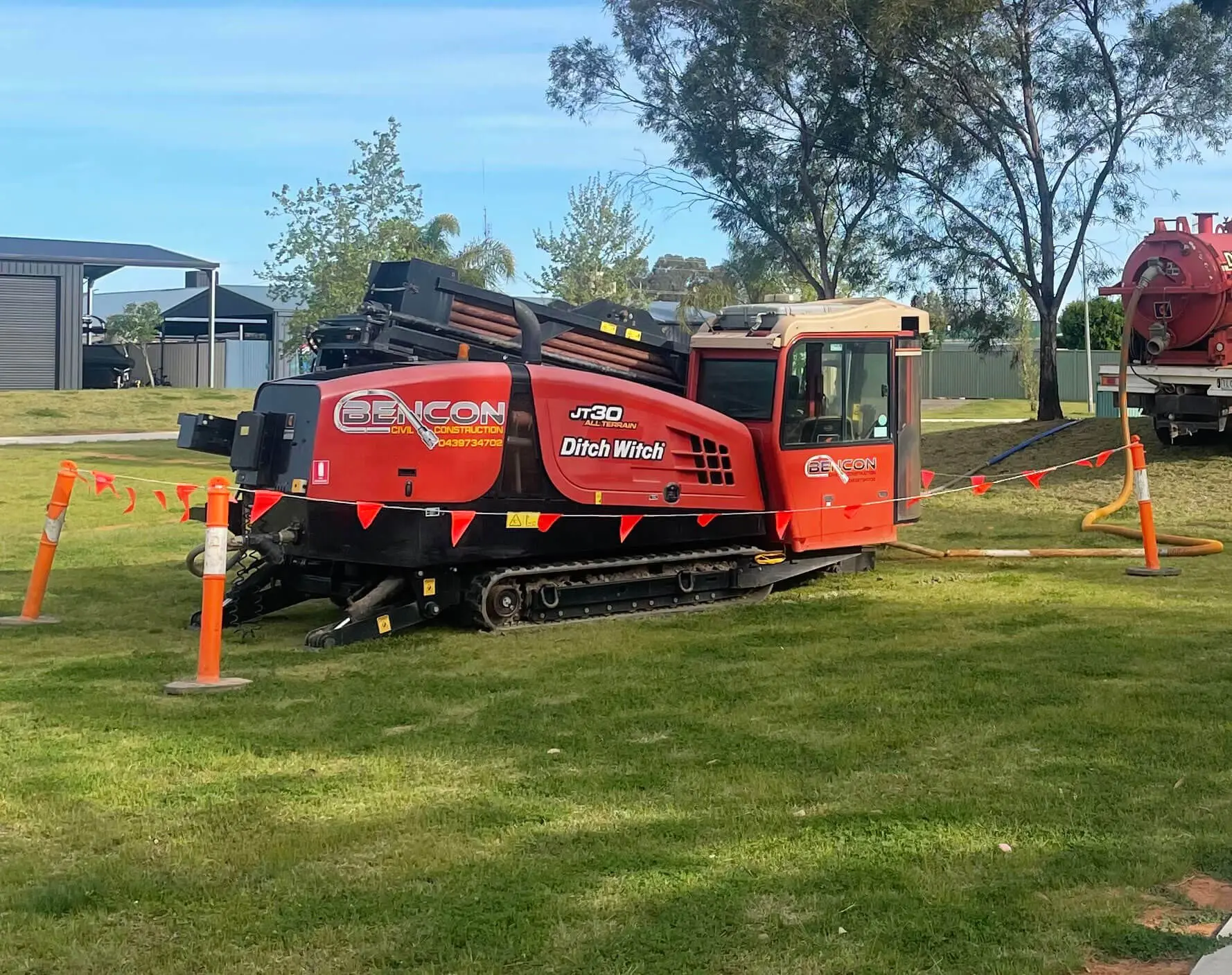 A red Ditch Witch J30 drilling machine, operated by a civil construction company, is set up on a grassy field surrounded by orange safety cones and caution tape. Trees, fences, and buildings are visible in the background.